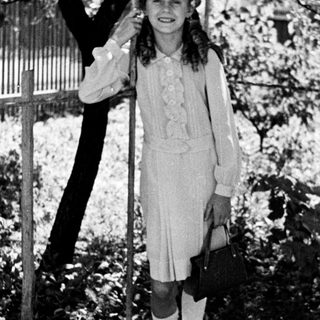 Here's a brief summary of the image: Girl in vintage dress stands near a tree.
