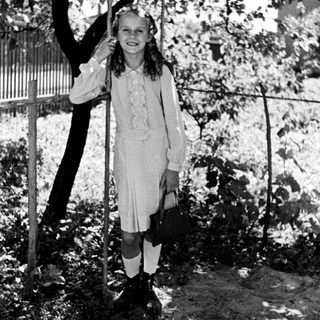 Here's a brief summary of the image: Girl in vintage dress stands near a tree.