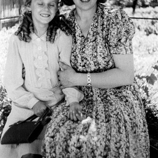 Two women in vintage dresses stand smiling outdoors.