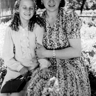 Two women in vintage dresses stand smiling outdoors.