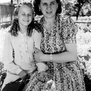 Two women in vintage dresses stand smiling outdoors.