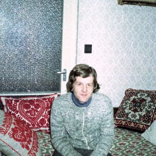 Man seated on a patterned couch in a vintage room.