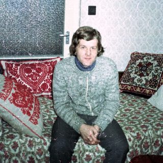 Man seated on a patterned couch in a vintage room.