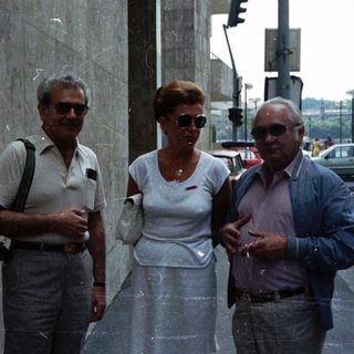 Three people standing on a street near a wall.