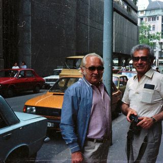 Two men standing on a street with cars and buildings.