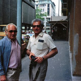 Two men standing on a street with cars and buildings.