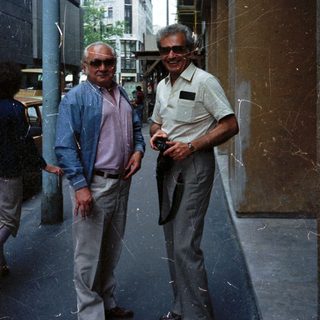 Two men in sunglasses stand on a city street.