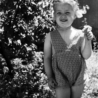 Girl smiles, stands near tree in vintage black and white photo.