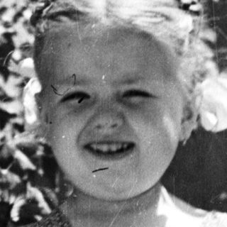Girl smiles, stands near tree in vintage black and white photo.