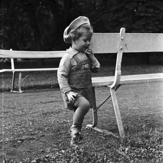 Here's a brief summary of the image: Young child stands near a bench in a grassy area.