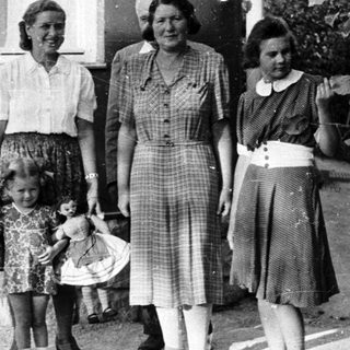 Girl with doll stands with three women outside a house.