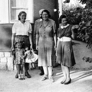 Girl with doll stands with three women outside a house.