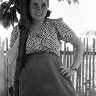 Woman stands smiling near a tree, in a vintage photograph.