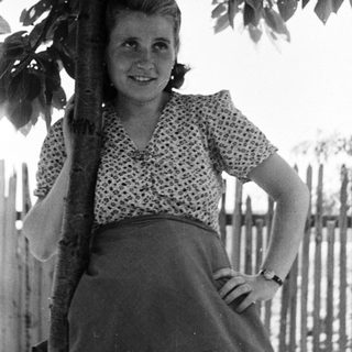 Woman stands smiling near a tree, in a vintage photograph.