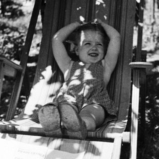Here's a brief summary of the image: Child sits in a chair, hands behind head, smiling.