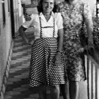 Two women stand on a tiled balcony, partially shaded.