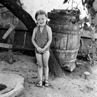Young girl stands near a barrel in a rustic setting.