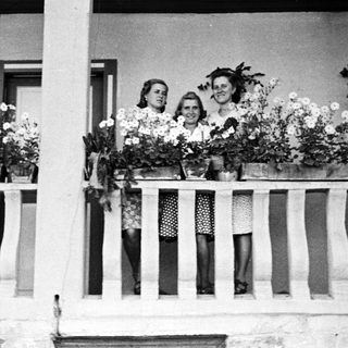 Three women stand on a balcony with flower boxes.