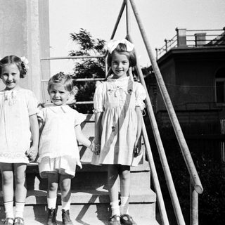 Three young girls stand on stairs in vintage clothing.