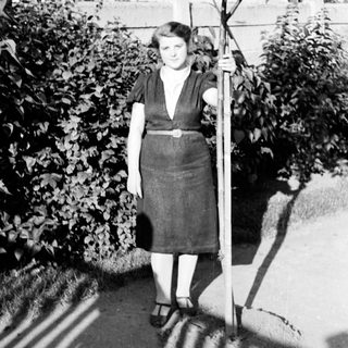 Woman stands holding a small tree in a garden.