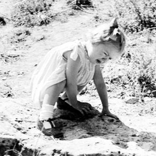 Here's a brief summary of the image: Young child kneeling in grass, examining the ground.