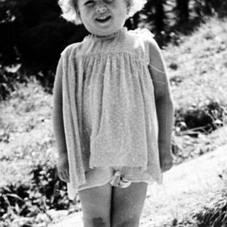 Here's a brief summary of the image: Young child stands on rocks, wearing a dress and shoes.
