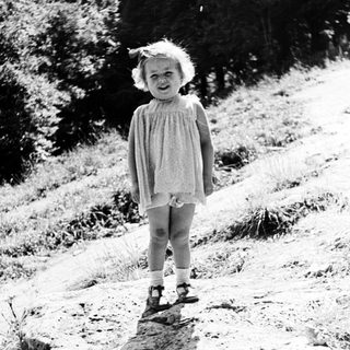Here's a brief summary of the image: Young child stands on rocks, wearing a dress and shoes.