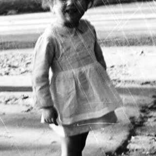 Here's a brief summary of the image: Young child stands outdoors in a vintage dress and boots.