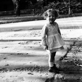 Here's a brief summary of the image: Young child stands outdoors in a vintage dress and boots.