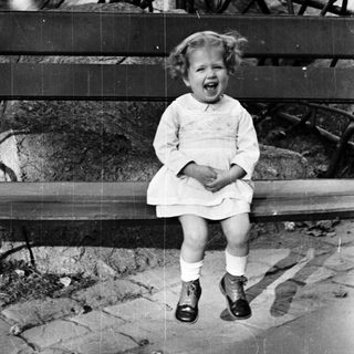 Here's a brief summary of the image: Young girl in a dress sits on a bench.