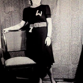 Woman in dress stands near a chair, posing for photo.