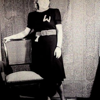 Woman in dress stands near a chair, posing for photo.