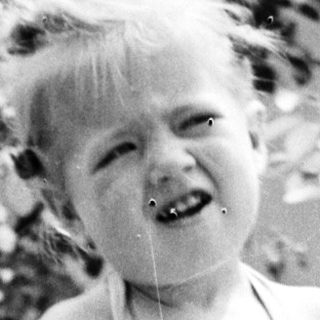 Here's a brief summary of the image: Child in overalls stands near foliage, smiling for the camera.