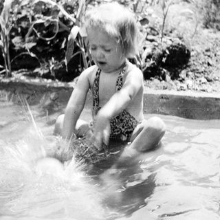 Child splashes in water, being held by an adult.