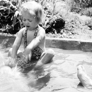 Child splashes in water, being held by an adult.