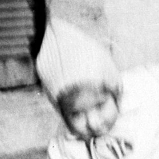 Here's a brief summary of the image: Child in a suit crouches near a building.