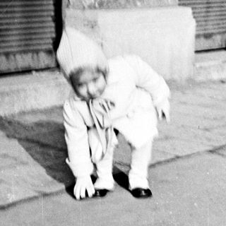 Here's a brief summary of the image: Child in a suit crouches near a building.