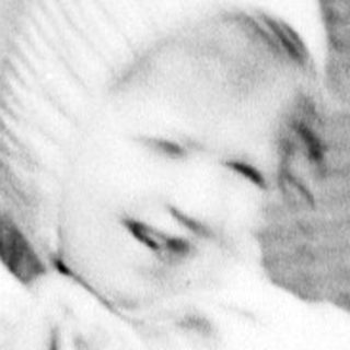 Here's a brief summary of the image: Baby in a coat stands next to a shuttered door.