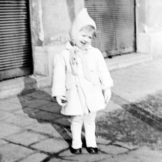 Here's a brief summary of the image: Baby in a coat stands next to a shuttered door.