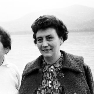 Two smiling women stand outdoors with a body of water behind.