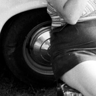 Woman sits near a car, applying cream to her face.