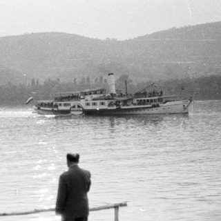 Three figures view a boat across a body of water.