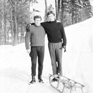 Here's a brief summary of the image: Two men stand with a sled in a snowy, wooded area.