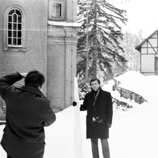 Man photographed in snowy landscape with buildings in background.