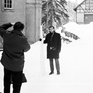 Man photographed in snowy landscape with buildings in background.