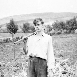 Young man stands in a field, holding a tool.