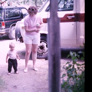 A woman and young child walking outdoors near parked vehicles.