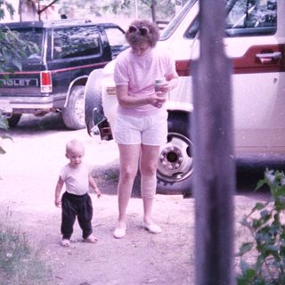 A woman and young child walking outdoors near parked vehicles.