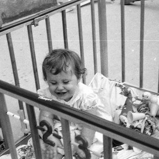 Baby smiling inside gated crib with stuffed toys.