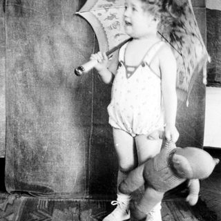 Young child holding umbrella indoors, dressed in diaper and overalls.
